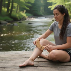 May include: A wooden massage tool is held near a person's leg. The tool is light brown and curved. The person is wearing a gray t-shirt and black shorts, sitting on a wooden dock near a river.