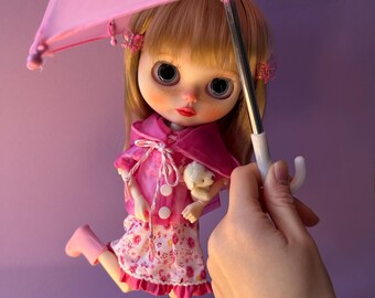 May include: A Blythe doll, dressed in a pink outfit and boots, holds a matching pink umbrella. The doll has blonde hair, large eyes, and is holding a small teddy bear. The doll is standing on a flower-shaped stand.