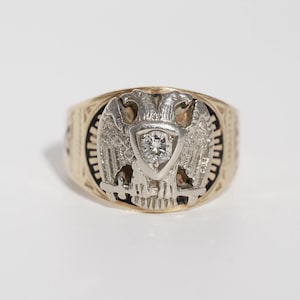 Puede incluir: Un anillo de oro con un diseño detallado con un emblema de águila bicéfala. El anillo tiene un diamante central engastado en un metal blanco, y la banda tiene un diseño estampado. El anillo es probablemente una pieza vintage o antigua.