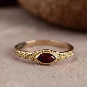 May include: A gold-colored ring with a maroon-colored, oval-shaped gemstone at the center. The ring band features an ornate, filigree design. The ring is set on a neutral-colored surface.