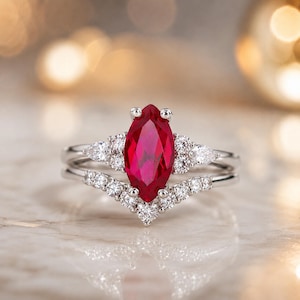 May include: A silver-toned ring set featuring a central marquise-cut red gemstone flanked by small round and pear-shaped clear stones. The design includes a curved band adorned with a row of round clear stones that nestles against the main ring.