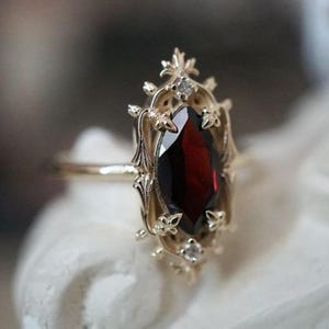 Könnte beinhalten: Ein goldener Ring mit einem zentralen, markisengeschliffenen, tiefroten Granat. Die Fassung ist mit aufwendigen Metallarbeiten verziert, darunter kleine Fleur-de-Lis-Akzente und zwei winzige, klare, rund geschliffene Steine an den Enden.