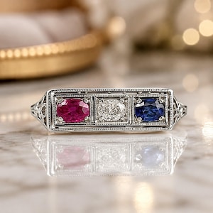 May include: A silver-toned ring featuring a rectangular setting with three oval-cut stones arranged horizontally. The center stone is clear, flanked by a vibrant red stone on the left and a deep blue stone on the right, all set in a detailed metal frame.