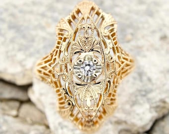May include: A gold filigree ring with a central, round, clear gemstone. The ring has an ornate, openwork design with floral and geometric patterns. The ring is set against a neutral, textured background.