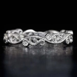 May include: A silver ring with an intricate filigree design. The ring features a repeating pattern of swirling shapes and small, round diamonds. The ring is set against a dark background, highlighting its delicate details and craftsmanship.