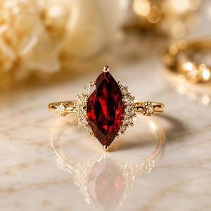 May include: A gold ring featuring a large marquise-cut deep red garnet gemstone at the center, surrounded by a halo of small clear round diamonds. The thin gold band includes additional small clear diamonds set on each side of the central stone.