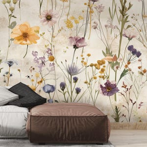 Puede incluir: Un papel pintado floral en acuarela con una variedad de flores silvestres en tonos naranja, morado, amarillo y rosa. Las flores se colocan sobre un fondo neutro y texturizado. Un puf marr&oacute;n y almohadas en primer plano.