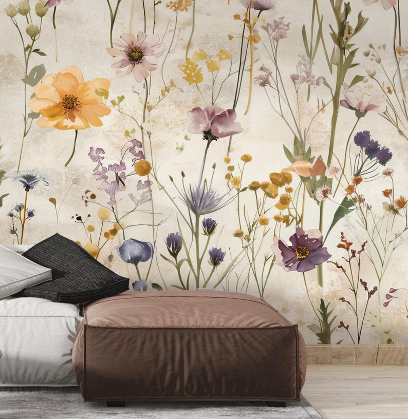 Puede incluir: Un papel pintado floral en acuarela con una variedad de flores silvestres en tonos naranja, morado, amarillo y rosa. Las flores se colocan sobre un fondo neutro y texturizado. Un puf marr&oacute;n y almohadas en primer plano.