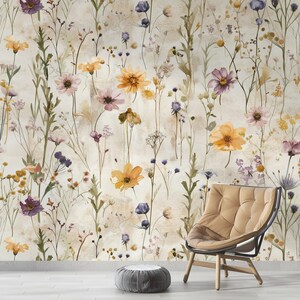 Puede incluir: Dise&ntilde;o de papel pintado con un patr&oacute;n floral en estilo acuarela. Las flores son en tonos amarillos, morados y rosas, sobre un fondo crema. Tambi&eacute;n se ve una silla beige y un puf gris.