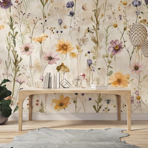 Puede incluir: Una mesa de madera clara frente a un papel pintado floral con flores de acuarela en tonos amarillos, morados y rosas. Una planta en maceta y libros est&aacute;n sobre la mesa. Una alfombra gris est&aacute; en el suelo.