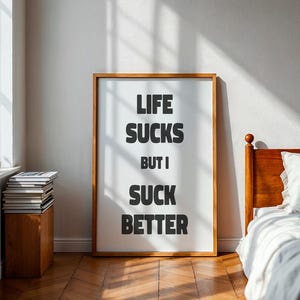 Puede incluir: Un cuadro enmarcado con el texto "LIFE SUCKS BUT I SUCK BETTER" en letras negras en negrita. El marco es de color marrón claro. El cuadro se exhibe en una habitación con suelo de madera y una cama.