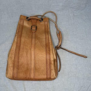 May include: A tan leather drawstring backpack featuring vertical stitched panels, a central buckle closure, and a single adjustable shoulder strap with metal hardware. The leather has a weathered texture with visible natural variations in color.