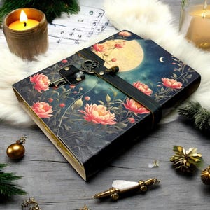 Puede incluir: Un diario decorativo con un diseño floral y lunar. La portada presenta una escena nocturna con una luna llena y flores rosas. Una correa de cuero negro y un cierre de llave aseguran el diario. Velas y objetos decorativos rodean el diario.