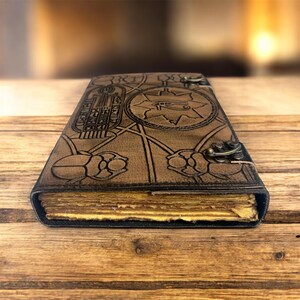 Puede incluir: Un diario de cuero marr&oacute;n con un dise&ntilde;o egipcio en relieve. La portada presenta jerogl&iacute;ficos, un s&iacute;mbolo de ojo y escarabajos. El diario tiene un cierre de metal y p&aacute;ginas amarillentas, lo que sugiere una est&eacute;tica vintage o antigua.