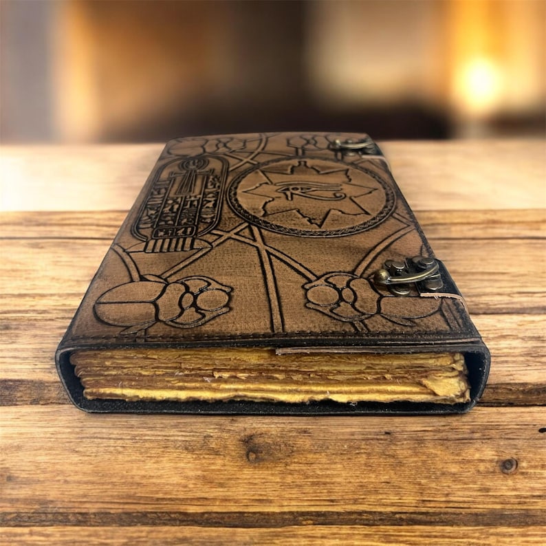 Puede incluir: Un diario de cuero marr&oacute;n con un dise&ntilde;o egipcio en relieve. La portada presenta jerogl&iacute;ficos, un s&iacute;mbolo de ojo y escarabajos. El diario tiene un cierre de metal y p&aacute;ginas amarillentas, lo que sugiere una est&eacute;tica vintage o antigua.