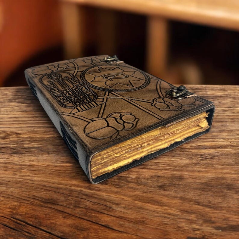 Puede incluir: Un diario de cuero marr&oacute;n con intrincados dise&ntilde;os en relieve, incluyendo jerogl&iacute;ficos y patrones florales. El diario tiene un cierre de broche y p&aacute;ginas amarillentas, lo que sugiere un estilo vintage. Descansa sobre una superficie de madera.