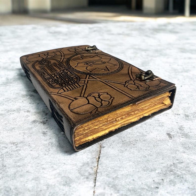 Puede incluir: Un diario de cuero marr&oacute;n con un dise&ntilde;o en relieve que presenta s&iacute;mbolos egipcios y motivos florales. El diario tiene un cierre de metal y p&aacute;ginas amarillentas envejecidas, lo que sugiere un estilo vintage o antiguo. La cubierta est&aacute; intrincadamente detallada.
