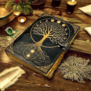 Puede incluir: Un diario negro con un diseño dorado de árbol de la vida en la portada. El árbol está rodeado por un círculo con lunas crecientes. El diario tiene un cierre de metal y páginas doradas. Descansa sobre una superficie de madera.