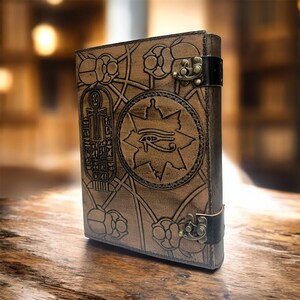 Puede incluir: Un diario de cuero marr&oacute;n con un dise&ntilde;o egipcio en relieve. La portada presenta jerogl&iacute;ficos, un s&iacute;mbolo del Ojo de Horus y patrones florales. Tiene un cierre de metal y una correa de cuero negro.