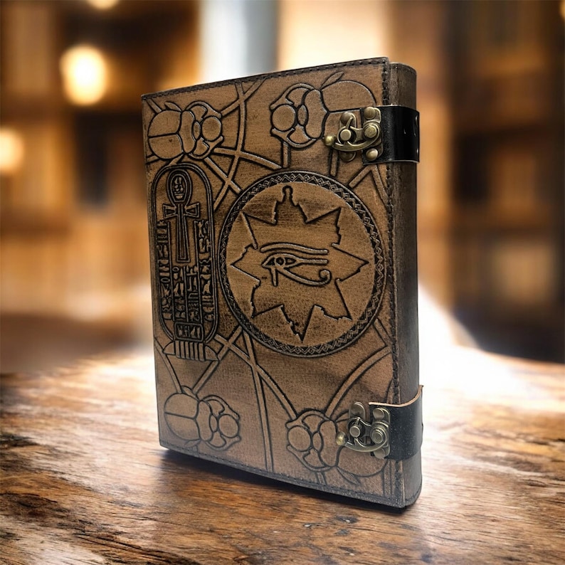 Puede incluir: Un diario de cuero marr&oacute;n con un dise&ntilde;o egipcio en relieve. La portada presenta jerogl&iacute;ficos, un s&iacute;mbolo del Ojo de Horus y patrones florales. Tiene un cierre de metal y una correa de cuero negro.