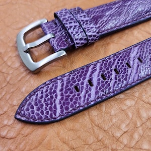 Puede incluir: Primer plano de una correa de reloj morada con un patrón texturizado, probablemente de cuero de avestruz. La correa tiene una hebilla plateada y está colocada sobre una superficie de cuero marrón. La correa tiene varios agujeros para ajustar.