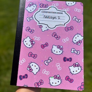 May include: Pink composition book with a pattern of Hello Kitty faces and bows. The cover reads "COMPOSITION BOOK" and has the name "Addisyn S." written in black. The book has a black spine.