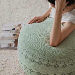 Pode incluir: Um pufe redondo em verde sálvia com uma superfície texturizada de ondas e uma faixa decorativa de motivos florais e linhas festonadas bordadas na lateral. O pufe possui um revestimento de tecido macio e um formato cilíndrico.