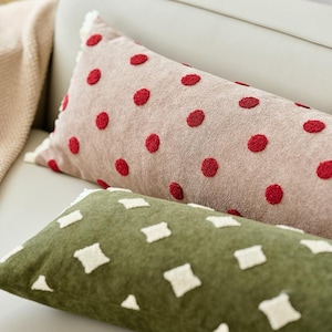 Peut inclure: Un coussin rectangulaire beige avec un motif à pois rouges texturés et un bord à franges blanches, placé derrière un coussin rectangulaire vert olive avec un motif de losanges blancs brodés et un bord à franges blanches.