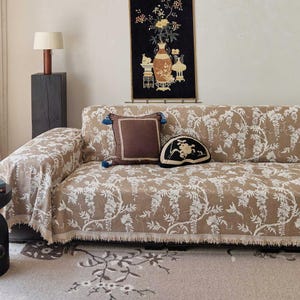 May include: A brown sofa with a white floral pattern is covered with a matching throw. A brown pillow with blue tassels and a black decorative pillow sit on the sofa. A rug with a floral design is on the floor.