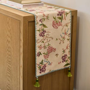 Op de afbeelding: Bloemen tafelloper met een beige achtergrond en een patroon van paarse en roze bloemen, groene bladeren en kleine blauwe accenten. De randen zijn afgewerkt met een lichtblauwe zigzagsteek en groene kwastjes.