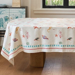 Può includere: Una tovaglia bianca con un motivo colorato di vita marina, tra cui cavallucci marini, stelle marine e tartarughe. Il bordo presenta un design a strisce blu e rosse. La tovaglia copre un tavolo di legno, creando un tema costiero.