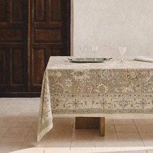 May include: A rectangular tablecloth featuring a beige, brown, and blue intricate pattern with floral and geometric motifs. The fabric drapes over a table surface, topped with a silver rectangular tray and three clear glass goblets.