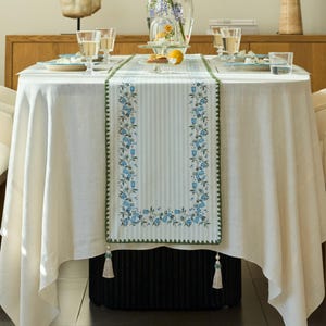 May include: A white tablecloth with a blue and green floral table runner. The runner has a striped background with blue floral designs and green trim. The table is set with glasses and plates.