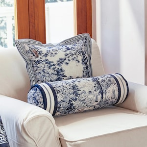 May include: Two decorative pillows on a white armchair. One is square, the other is a bolster. Both feature a blue and white toile design with a nature scene. The square pillow has a border and a small animal in the design.