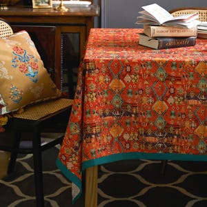 May include: A vibrant orange tablecloth with a geometric pattern in shades of yellow, blue, and purple. The tablecloth has a teal border and is draped over a wooden table. A decorative pillow with floral designs rests on a chair.