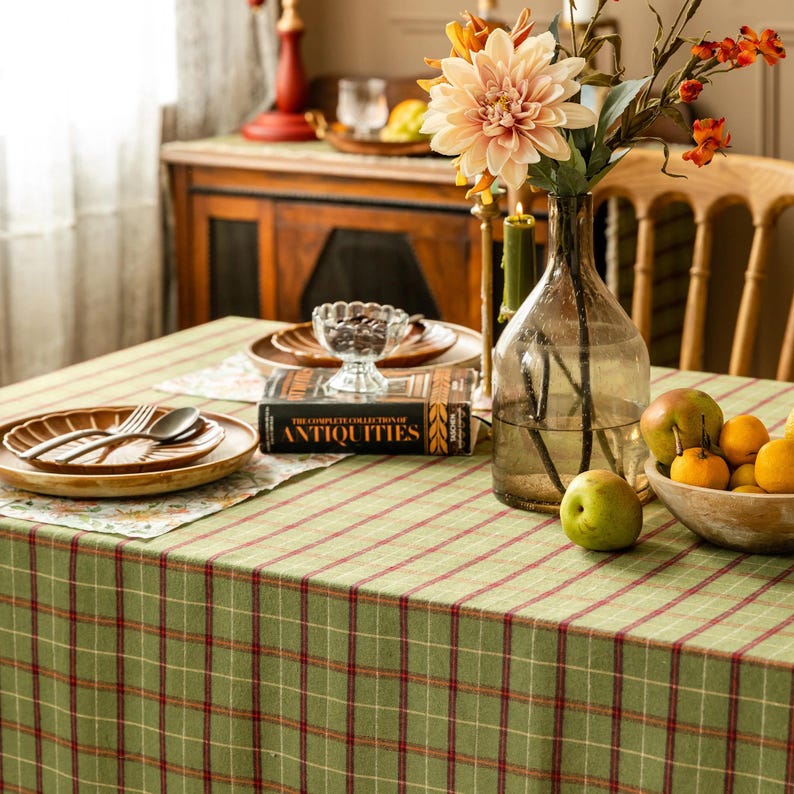 May include: A dining table set with a green and red plaid tablecloth. The table is set with wooden plates, silverware, a glass vase with flowers, and a bowl of fruit. A book titled "ANTIQUITIES" is also on the table.