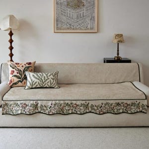 May include: A beige sofa cover with a scalloped edge featuring a detailed botanical and bird print in green, brown, and red tones. The cover is draped over a light-colored sofa, accompanied by a square floral pillow and a rectangular leaf-patterned pillow.