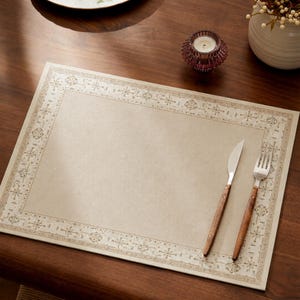May include: A rectangular beige placemat featuring a decorative border with a repeating star and floral pattern in a darker shade. A stainless steel knife and a fork with brown wooden handles are placed on the right side of the placemat.