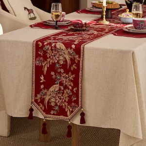 May include: A red table runner with a floral and bird pattern, edged with a beige border and tassels. It's laid on a beige tablecloth, with a dining setting including plates, glasses, and a decorative pillow in the background.