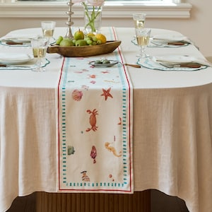 May include: A dining table set for a meal, featuring a white tablecloth and a decorative runner. The runner has a cream background with red and teal borders and sea creature illustrations. The table is set with plates, glasses, and silverware.