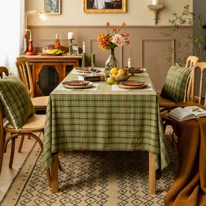 Puede incluir: Una mesa de comedor preparada para una comida, con un mantel a cuadros verdes con detalles rojos. La mesa está puesta con platos de madera, manteles individuales y un jarrón con flores. Dos cojines a cuadros verdes y sillas de madera completan la escena.