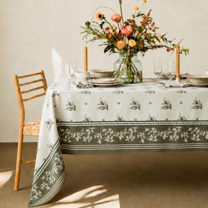 May include: A rectangular tablecloth featuring a cream-colored center with a repeating olive branch pattern and a wide, dark olive green border with a detailed floral vine print. The cloth is draped over a table set with plates and a flower vase.