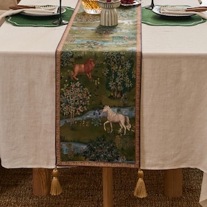 May include: A rectangular table runner featuring a green background with a detailed print of a lion, a white horse, trees, and flowers. The runner has a dark border and two gold-colored tassels hanging from the bottom corners.