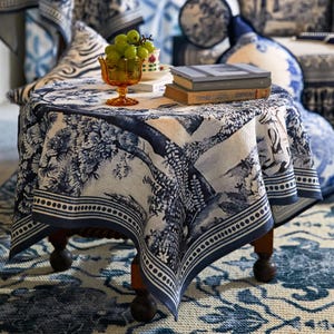 May include: A small, round wooden table covered with a blue and white patterned tablecloth. The tablecloth has a detailed landscape design. Books, a glass bowl of green grapes, and a teacup sit on the table. The table is on a patterned rug.