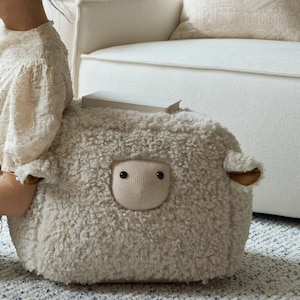 May include: A cream-colored, plush, sheep-shaped ottoman featuring a textured, curly fleece exterior, a smooth, light beige fabric face with two small black eyes, and two small ears with brown inner accents, topped with a rectangular white book.