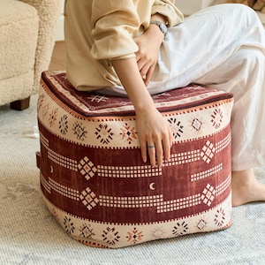 Könnte beinhalten: Ein quadratischer Boden-Pouf in den Farben Dunkelrot und Creme mit einem sich wiederholenden geometrischen Muster. Das Design zeigt horizontale Bänder aus kleinen Quadraten, Rautenformen und Mondsichel-Symbolen auf der Stoffoberfläche.