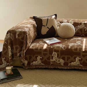 Può includere: Una coperta da divano marrone con un motivo ripetuto di cavalli e geometrico. Un cuscino abbinato con un motivo a testa di cavallo e un cuscino decorativo rotondo color crema sono sul divano. Sono visibili anche un piccolo libro e una candela.