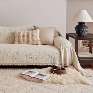 May include: A cream-colored textured sofa throw blanket with fringe edges, featuring a subtle speckled pattern. The blanket is draped over a sofa with a rectangular faux fur pillow and a square beige pillow, alongside a dark side table with a lamp.