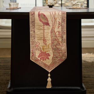 Puede incluir: Un camino de mesa rectangular de tela beige tejida con un diseño estampado de un flamenco rosa junto a follaje estilizado. El camino tiene un extremo en punta rematado con una borla decorativa de color crema y un borde oscuro.