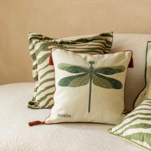 May include: A square throw pillow featuring a green dragonfly graphic and the name Amelia printed in green. The pillow has dark red piping and tassels at the corners. Behind it sits a second pillow with a green and white zebra stripe pattern.